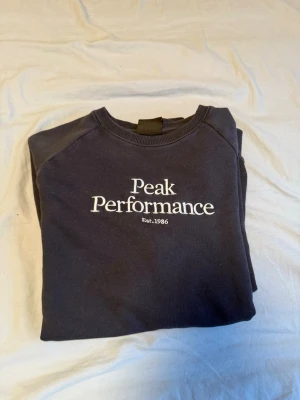 Mörkblå sweatshirt Peak Performance - Mörkblå sweatshirt från Peak Performance med vit logotyp och text 'Est. 1986' på bröstet. Klassisk rund halsringning, långa ärmar och ribbade muddar vid ärmslut och nederkant. Perfekt för en avslappnad och stilren look. Lite solblekt 