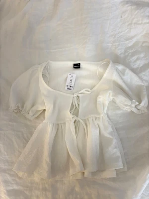 Vit topp från Gina Tricot - Supersöt vit topp från Gina Tricot i storlek XS. Toppen har puffiga korta ärmar, knyt framtill och lätt rynkad struktur i tyget. Perfekt för dig som gillar romantisk och luftig stil. Materialet känns mjukt och lätt. Säljs inte längre på Gina.