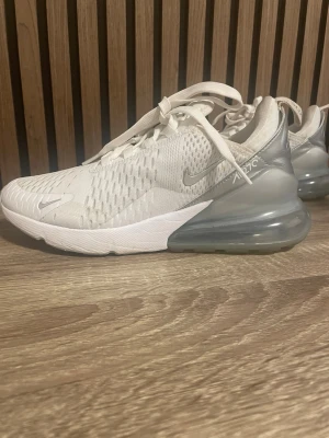 Nike Air Max 270 vita sneakers - Nike Air Max 270 sneakers i vitt med grå detaljer och stor synlig luftbubbla i hälen. Ovandelen är i mesh för bra ventilation och har snörning. Swoosh-logga på sidan och 270-tryck. Perfekta för en sportig och modern look.