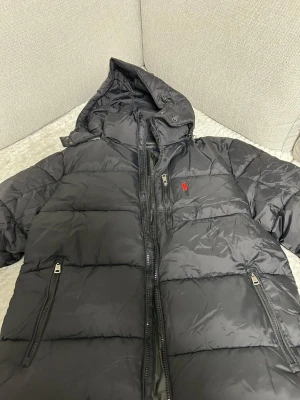 Svart dunjacka från Ralph Lauren - Snygg svart dunjacka från Ralph Lauren med huva och två dragkedjeförsedda fickor. Jackan har quiltad design och den klassiska röda loggan på bröstet. Perfekt för kalla dagar och riktigt stilren look.