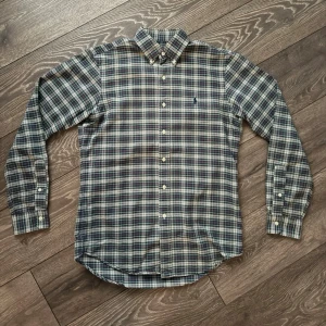 Rutig blå skjorta med knappar - Klassisk rutig skjorta i blått, vitt och grått med button-down krage och långa ärmar. Skjortan har knäppning framtill och en liten broderad logga på bröstet. Perfekt för en avslappnad men ändå stilren look.