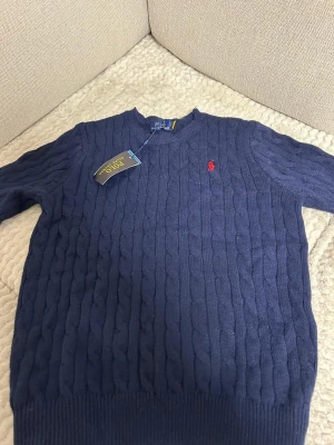 Mörkblå kabelstickad tröja Polo Ralph Lauren - Kabelstickad mörkblå tröja från Polo Ralph Lauren med rund halsringning och röd broderad logga på bröstet. Tröjan har ribbade muddar vid ärmslut och nederkant, vilket ger en klassisk och stilren look. Perfekt för dig som gillar tidlös design.