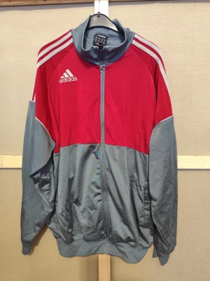 Adidas träningsjacka i rött och grått – retro och sportig - M - Sportig Adidas jacka i rött och grått med klassiska vita ränder på axlarna. Jackan har hel dragkedja framtill, hög krage och två sidofickor. Tillverkad i polyester och har långärmad design. Perfekt för träning eller chill.