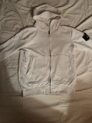 Vit hoodie från Stone Island - Säljer en clean vit hoodie från Stone Island med dragkedja framtill och klassisk logga på ärmen. Hoodien har två stora fickor fram och ribbade muddar. Perfekt för dig som gillar streetwear och vill ha något stilrent. Materialet känns mjukt och skönt.