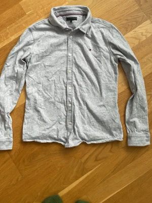 Ljusgrå skjorta från Tommy Hilfiger - Snygg ljusgrå skjorta från Tommy Hilfiger med klassisk krage och knappar framtill. Liten logga på bröstet och stor broderad logga på ryggen. Tillverkad i mjuk bomull och har långa ärmar. Perfekt för en clean och stilren look.