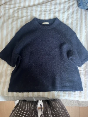 Mörkblå stickad tröja med kort ärm - Säljer en mörkblå stickad tröja med korta ärmar och rund hals. Tröjan har en avslappnad passform och är gjord i ett mjukt, luddigt material som känns skönt mot huden. Perfekt för lager på lager och enkel att styla till olika outfits.