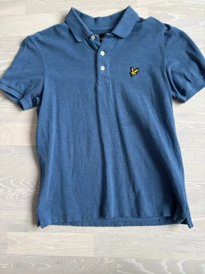 Blå pikétröja från Lyle & Scott - Klassisk blå pikétröja från Lyle & Scott med gul broderad logga på bröstet. Tröjan har krage, knäppning med tre vita knappar och korta ärmar. Perfekt till jeans eller shorts för en clean och enkel stil.
