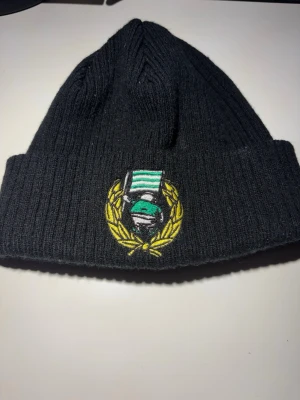 Svart ribbstickad mössa med brodyr - Säljer en svart ribbstickad Bajen mössa med uppvikt kant och en broderad patch framtill som visar en grön och vit keps omgiven av en gul lagerkrans. Perfekt för dig som vill ha en stilren men ändå unik accessoar till vintern.