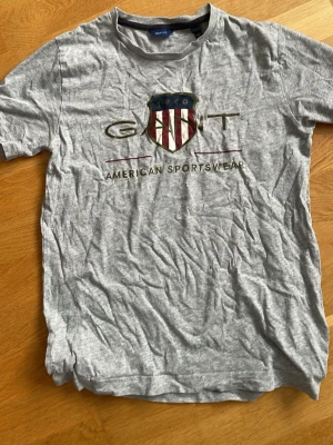 Grå GANT t-shirt med tryck - Snygg grå t-shirt från GANT med stort tryck framtill som visar ett amerikanskt inspirerat emblem och texten 'American Sportswear'. Klassisk rund hals och korta ärmar. Tillverkad i mjuk bomull för skön känsla.