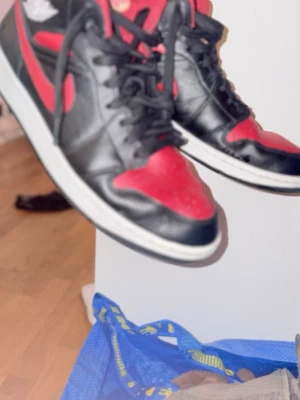 Nike Air Jordan 1 svart/röd sneakers - Säljer ett par klassiska Nike Air Jordan 1 sneakers i svart och rött skinn med vita sulor. Ikonisk design med snörning, rund tå och Air Jordan-logga på sidan. Perfekta för dig som gillar streetwear och vill sticka ut med en tidlös modell.