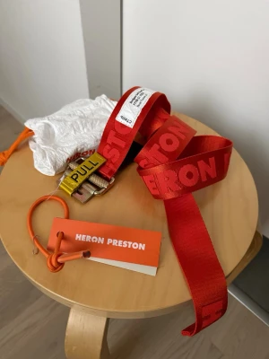 Rött bälte från Heron Preston - Säljer ett rött statement-bälte från Heron Preston med tydlig logga längs hela bandet. Bältet har en robust metallspänne med gul 'PULL'-detalj och kommer med originalpåse och tagg. Perfekt accessoar för att lyfta din outfit. Oanvänt, prislappen sitter kvar. Kommer med dustbag.