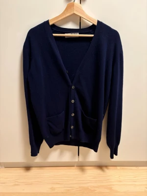 Mörkblå cardigan från Alan Paine - Klassisk mörkblå cardigan i 100% geelong lammull från Alan Paine. Tröjan har v-ringning, knäppning framtill med fem knappar och två fickor. Perfekt för lager på lager och en stilren look.