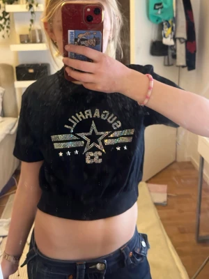 Svart croppad t-shirt med stjärndetalj - Säljer en svart croppad t-shirt från SOL'S med glittrande stjärndetalj och texten 'GUARDIAN' framtill. T-shirten har korta ärmar och coola paljetter i flera färger som ger en unik look. Perfekt för dig som vill sticka ut lite extra.