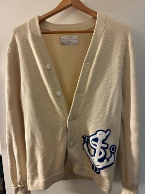 Beige kofta från These Glory Days, Cardigan  - Snygg beige kofta från These Glory Days med knäppning framtill och ett stort broderat märke i vitt och blått vid höften. Koftan har ribbade muddar och en klassisk passform, perfekt att slänga över en t-shirt.