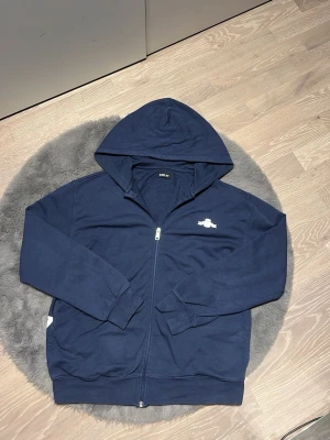 Marinblå hoodie från Replay - Snygg marinblå hoodie från Replay med dragkedja framtill och stor huva. Framsidan har en liten vit Replay-logga på bröstet och baksidan pryds av ett stort vitt REPLAY-tryck längs nederkanten. Tillverkad i mjuk bomull, perfekt för en avslappnad stil.