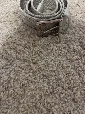 Ljusgrått flätat bälte med silverdetaljer - Snyggt ljusgrått bälte med flätad design och silverfärgat spänne. Bältet har detaljer i konstläder vid ändarna och runt spännet. Passar perfekt till jeans eller chinos för en clean och stilren look.