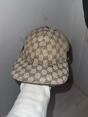 Gucci beige keps  - Säljer en beige keps från Gucci med det klassiska GG-monogrammet över hela kepsen. På sidan finns en grön och röd rand som ger extra lyxig känsla. Kepsen har böjd skärm och justerbar rem bak. Materialet är canvasliknande och känns riktigt premium. Den är i storlek xs en ovanlig storlek som är svårare att hitta jämfört med de andra storlekarna, Äkta, det är den gamla model som är nu sällsynt och svårt att köpa samt i bra skick, bara hör av er angående frågor!! Pris går och diskutera!