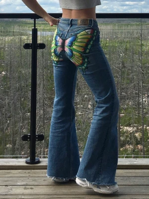 Blå bootcut jeans med fjärilsmålning - Unika handmålade bootcut jeans med stor färgglad fjäril bak. Byxorna har en snygg passform med flare, råa kanter och medelhög midja. De sitter bekvämt och tyget känns mjukt och skönt och går att röra sig i. Fjärilsmålningen är gjord i textilfärg och är fixerad för att hålla. Vid tvätt: vänd plagget ut och in och tvätta på milt program. Storlek: 28/33 Midjemått: 79cm, Innerbenslängd: 83cm