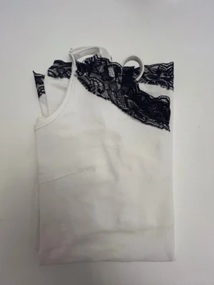 Vit linnetopp med marinblå spetskant - Snygg vit linnetopp med tunna axelband och dekorativ svart spetskant upptill och nedtill. Perfekt för att addera en feminin touch till din outfit. Materialet känns mjukt och lätt mot huden.