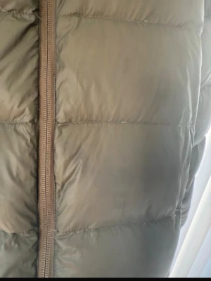 Grön dunjacka från Sail Racing - Mörkgrön dunjacka från Sail Racing med huva och dragkedja framtill. Jackan har quiltad design och logga på bröstet. Perfekt för kalla dagar och har en klassisk pufferlook som är både snygg och funktionell.