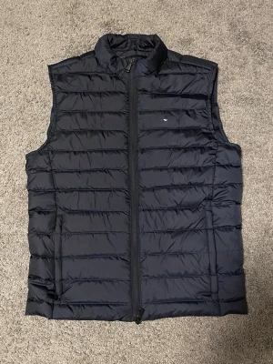 Svart dunväst från J.Lindeberg - Snygg svart dunväst från J.Lindeberg med quiltad design och hög krage. Västen har dragkedja framtill och två sidofickor. Endast testad, men säljs då den är för liten. Strl. XS                                                                                 Nypris: 1100:-