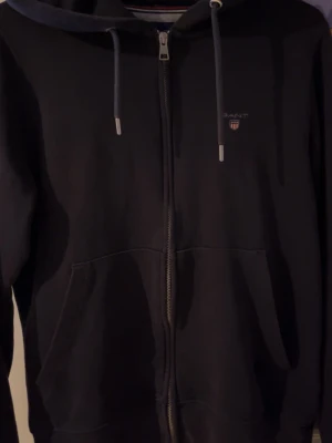 Svart zip hoodie från Gant - Snygg svart hoodie från Gant med dragkedja framtill och justerbar huva med snören. Klassisk Gant-logga broderad på bröstet och två stora fickor framtill. Tillverkad i mjuk bomullsblandning som är skön att ha på sig.