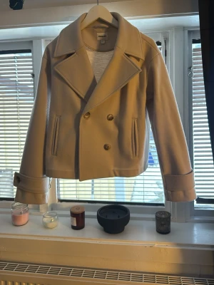 Beige kappa från Kappahl med knappar - Stilren beige kavaj från Kappahl med dubbelknäppning och breda slag. Kappan har långa ärmar, två fickor framtill och en croppad passform. Perfekt för dig som gillar klassisk men trendig stil.