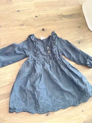 Grå klänning med volang och broderi - Söt grå klänning med långa ärmar, volangdetaljer längs knäppningen och broderade blommor vid fållen. Klänningen har två dekorativa knappar framtill och en vacker vågig kant nedtill. Perfekt för dig som gillar romantiska detaljer.