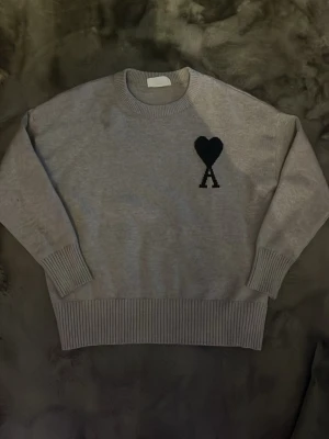 Grå stickad tröja från Ami - Säljer en stilren grå stickad tröja från Ami Alexandre Mattiussi med svart hjärtdetalj och A på bröstet. Tröjan har rund hals, ribbade muddar och bred ribbad nederkant. Perfekt för dig som gillar minimalistisk design och snygga detaljer.