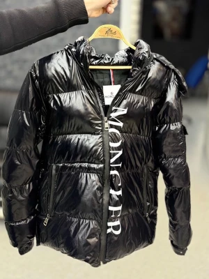 Svart dunjacka från Moncler - Snygg svart dunjacka från Moncler med glansig finish och stor vit logga längs dragkedjan. Jackan har hög krage, två sidofickor med dragkedja och quiltad design. Perfekt för kalla dagar när du vill hålla stilen.