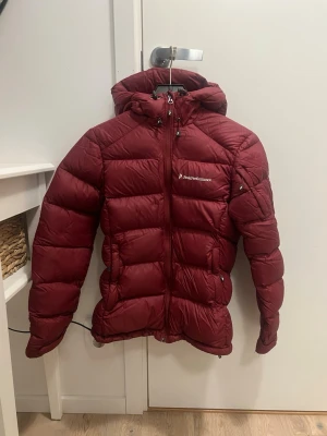 Vinröd dunjacka från Peak Performance - Snygg vinröd dunjacka från Peak Performance med huva och dragkedja framtill. Jackan har en quiltad design och två sidofickor med dragkedja samt ett ficka på armen med dragkedja. Perfekt för kalla dagar och har en modern, puffig look.