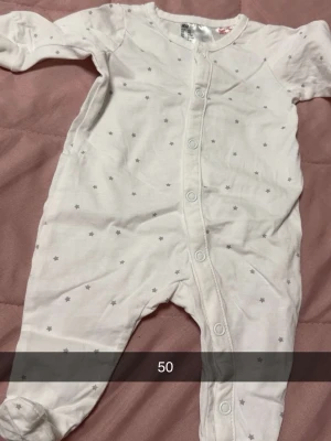 Vit sparkdräkt med små stjärnor - Mysig vit sparkdräkt med små grå stjärnor och tryckknappar framtill. Långärmad och med fötter, perfekt för att hålla bebisen varm. Tillverkad i mjukt bomullstyg för extra komfort.