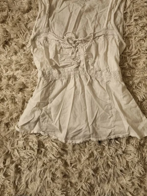 Vit linnetopp med snörning från Lindex - Söt vit linnetopp från Lindex med snörning framtill och spetsdetaljer vid axelbanden. Toppen har smock i midjan för en snygg passform och är ärmlös, perfekt för varma dagar. Skulle säga att den passar som en 38/lite större S.