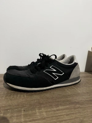 Svarta New Balance sneakers i mocka - Klassiska svarta New Balance sneakers med grå detaljer och vit logga på sidan. Skorna har snörning, rund tå och platt sula. Ovandelen är i mocka och textil, medan hälen är i grå mocka. Perfekta för en sportig och avslappnad stil.