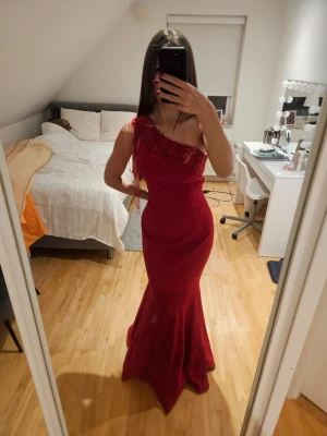 Röd långklänning med pärlor - Snygg röd långklänning med oneshoulder-design och pärldetaljer över axeln. Klänningen har en figurnära passform och är tillverkad i ett glansigt material som ger en elegant look. Perfekt för speciella tillfällen där du vill sticka ut.