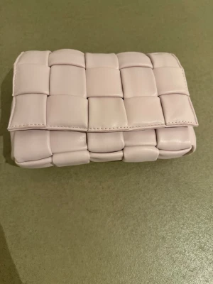 Ljus rosa flätad axelväska i skinn - Trendig ljus rosa axelväska med flätad design och justerbart axelband. Väskan har en mjuk, vadderad look och är tillverkad i skinnliknande material. Perfekt för dig som vill ha en stilren och modern accessoar till din outfit.