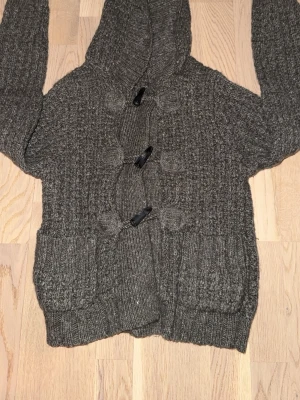 Stickad hoodie från Calvin Klein Jeans - Kan diskutera pris! Mysig stickad hoodie i mörkgrått från Calvin Klein Jeans. Tröjan har stora fickor framtill, ribbade muddar och snygga knappar. Perfekt för kyliga dagar och har en avslappnad, trendig look. 