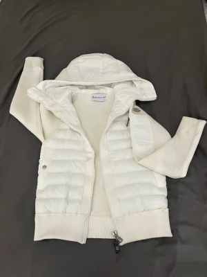 Vit cardigan jacka från Moncler - Vit cardigan jacka från Moncler med stickade ärmar och quiltad front. Jackan har huva, dragkedja framtill och en liten ficka med knapp på ena ärmen. Perfekt för lager-på-lager och snygg till många outfits.