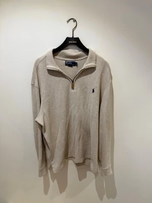 Beige halfzip från Polo Ralph Lauren - Säljer denna snygga beige halfzip-tröja från Polo Ralph Lauren med klassisk blå logga broderad på bröstet. Tröjan har krage med dragkedja och är tillverkad i mjukt bomullsmaterial. Perfekt för en clean och avslappnad stil. 