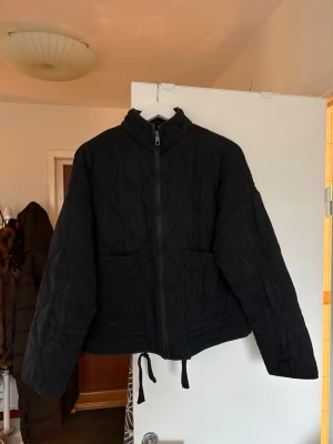 Svart quiltad pufferjacka från Zara - Säljer en svart quiltad pufferjacka från Zara med dragkedja framtill, två stora fickor och snörning i nederkant. Jackan har en loose passform och hög krage, perfekt för kyliga dagar. Enkel och stilren design som funkar till det mesta.