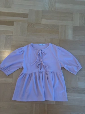 Rosa blus med knyt och puffärm - Supersöt rosa blus med två knytband framtill och puffiga trekvartsärmar. Blusen har en lös passform och är gjord i ett mjukt tyg som ger en avslappnad men trendig look. Perfekt att styla med jeans eller kjol för en cool vibe.