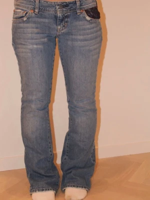 Blå bootcut jeans med spetsdetaljer - Säljer ett par blå bootcut jeans med unika bakfickor i mörk spets. Jeansen har klassisk femficksmodell, låg midja och är tillverkade i denim med stretch. Perfekta för dig som vill ha en cool twist på dina vardagsjeans. Inte mina bilder!! Storlek S-M