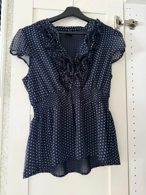 Mörkblå prickig blus med volang - Söt mörkblå blus med vita prickar och volangdetaljer längs V-ringningen. Blusen har korta ärmar i transparent material och smock i midjan för en snygg passform. Perfekt till jeans eller kjol för en trendig look.