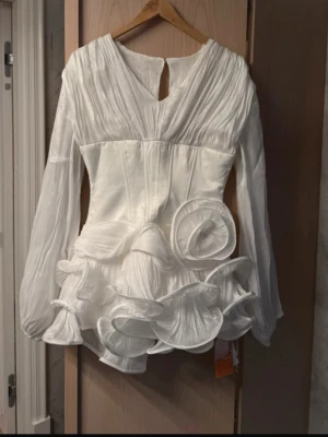 Vit kortklänning med volang och rosett - Supersnygg vit kortklänning med transparenta långärmar, markerad midja och stora volangdetaljer samt en dekorativ rosett framtill. Klänningen har en unik struktur och ger en riktigt statement-look. Perfekt för dig som vill sticka ut! Perfekt till studenten