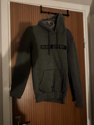 Grå hoodie från Philipp Plein - Snygg grå hoodie från Philipp Plein med svart logga framtill och matchande dragsnören i huvan. Hoodien har en stor magficka och ribbade muddar vid ärmar och nederkant. Perfekt för en chill och trendig look.