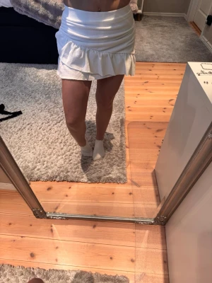 Vit kortkjol med volang - Supersöt vit kortkjol med volangkant och figurnära passform från Nelly . Kjolen har en lätt struktur i tyget och sitter högt i midjan. Perfekt för dig som gillar en feminin och trend sopad look.