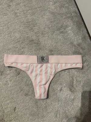 Rosa-vit randig trosa från Victoria's Secret - Säljer en söt stringtrosa från Victoria's Secret i rosa och vitt med klassiska ränder. Trosan har bred resår med glittrande VS-logga framtill och är tillverkad i mjukt material för extra komfort. Perfekt för dig som gillar en feminin och trendig stil.