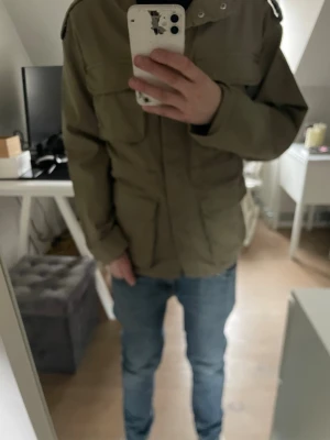 Olivgrön parkajacka med fickor - Säljer en olivgrön parkajacka med fyra stora fickor framtill och axelklaffar. Jackan har en rak passform och stängs med dragkedja och knappar. Perfekt för dig som gillar en enkel men stilren look.