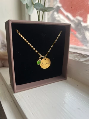 Guldigt halsband med grön sten - Snyggt halsband i guldfärgad metall med en rund bricka där det står 'Emma' ingraverat och ett litet hänge med en grön månads sten(Augusti), stenen går att plocka av om man vill de. Kedjan är tunn och elegant, perfekt för att ge en personlig touch till din stil. Säljer för att jag inte andvänder guld. Fått i present för nått år sedan, aldrig andvänd.
