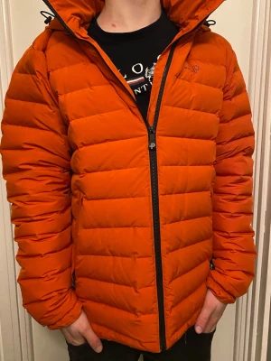 Orange dunjacka från Pelle P, Urbis Jacket, Cayenne  - Säljer en riktigt snygg orange dunjacka från Pelle P med huva och svart dragkedja. Jackan har quiltad design, lång ärm och logga på ärmen. Perfekt för kalla dagar och ger en cool look till vinterns outfits. Använd fem tillfällen, nypris 3000